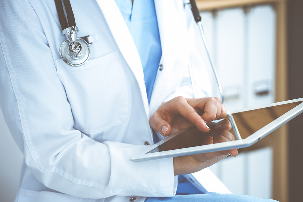 Woman doctor using white tablet computer while sitting at chair in hospital, close-up.
