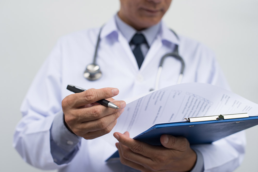 Male doctor on duty in white coat reading patient's information with pen in hand, filling prescription or checklist document, close up, selective focus on pen, health and medical concept.